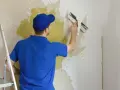 Mężczyzna w niebieskiej koszulce i czapce usuwa starą farbę ze ściany za pomocą szpachelki. Obok stoi drabina.