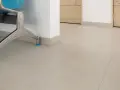 Jasnoszara podłoga z płytek, z widocznym cokołem o niewielkiej wysokości, przy metalowej nodze niebieskiego krzesła.