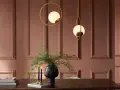 Elegancka jadalnia w odcieniu kolor ziemi. Dwa złote żyrandole w kształcie kół z mlecznymi kulami, czarny wazon z zieloną roślinnością i dwie świece na drewnianym stole.