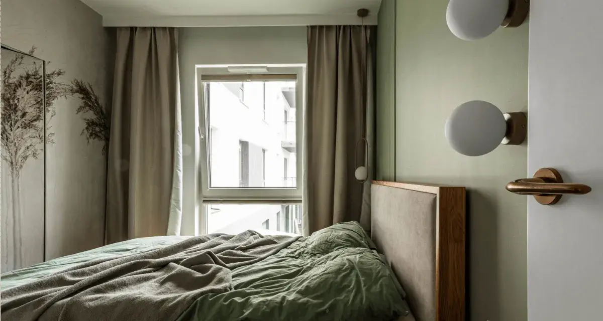 Przytulna, mała sypialnia z łóżkiem, zieloną pościelą i oknem z zasłonami. Dwie kule lamp na ścianie.