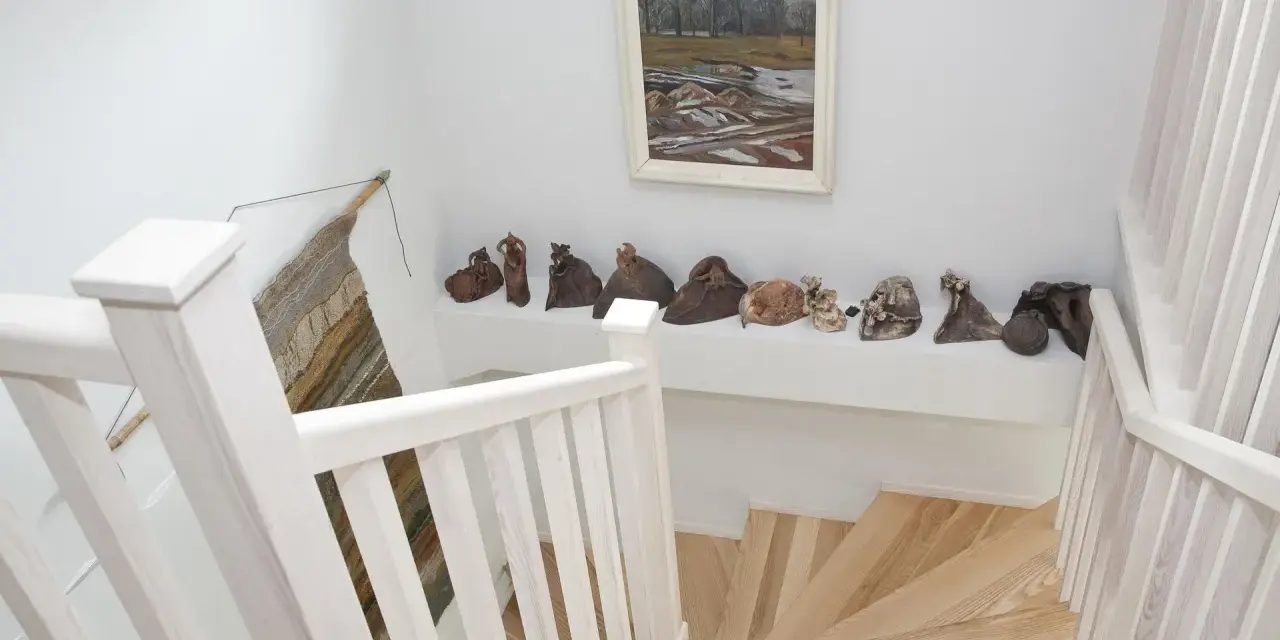 Jak wykończyć klatkę schodową w domu? Jasne drewno, białe balustrady i ciekawa ekspozycja rzeźb tworzą przytulną przestrzeń.