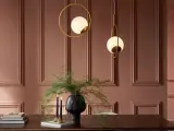 Elegancka jadalnia w odcieniu kolor ziemi. Dwa złote żyrandole w kształcie kół z mlecznymi kulami, czarny wazon z zieloną roślinnością i dwie świece na drewnianym stole.