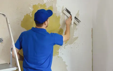 Mężczyzna w niebieskiej koszulce i czapce usuwa starą farbę ze ściany za pomocą szpachelki. Obok stoi drabina.