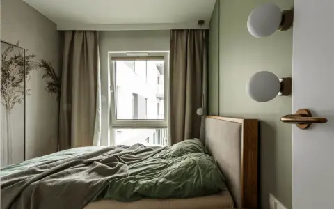 Przytulna, mała sypialnia z łóżkiem, zieloną pościelą i oknem z zasłonami. Dwie kule lamp na ścianie.