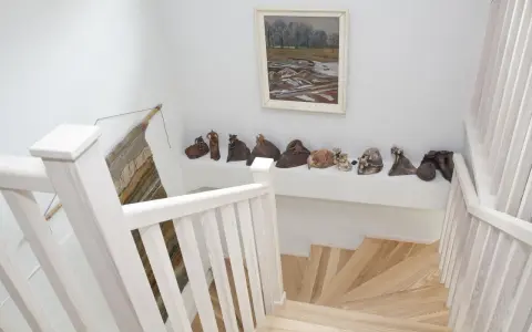 Jak wykończyć klatkę schodową w domu? Jasne drewno, białe balustrady i ciekawa ekspozycja rzeźb tworzą przytulną przestrzeń.