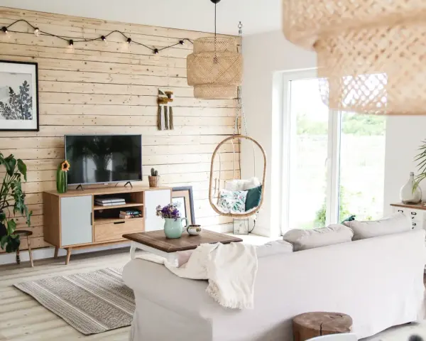 Przytulny salon z drewnianą ścianą, gdzie stara boazeria w nowej odsłonie tworzy ciepłą atmosferę. Wygodna sofa, wiszący fotel i rośliny dodają uroku.