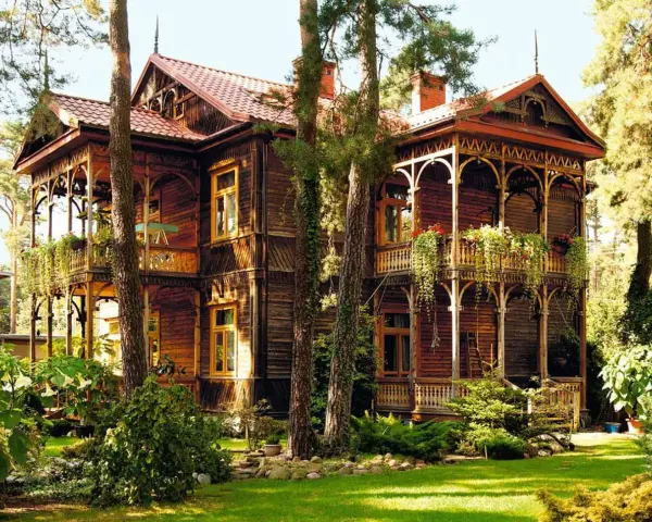 Drewniany dom w stylu świdermajer, z werandami i bogatymi zdobieniami, otoczony zielenią.
