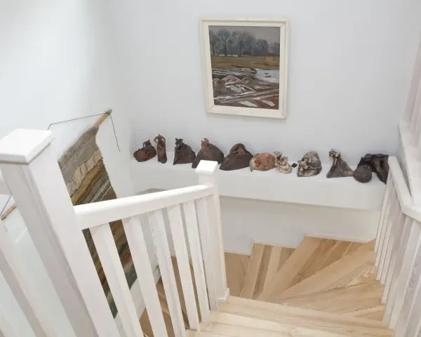 Jak wykończyć klatkę schodową w domu? Jasne drewno, białe balustrady i ciekawa ekspozycja rzeźb tworzą przytulną przestrzeń.