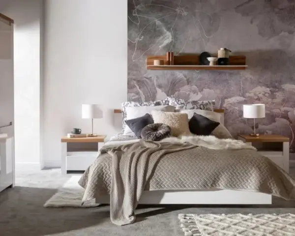 Przytulna sypialnia inspiracje: łóżko z miękkim kocem i ozdobnymi poduszkami, kwiatowa tapeta, drewniana półka, szafki nocne z lampkami.