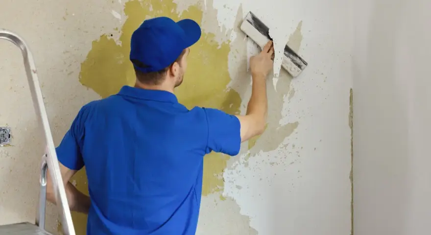 Mężczyzna w niebieskiej koszulce i czapce usuwa starą farbę ze ściany za pomocą szpachelki. Obok stoi drabina.