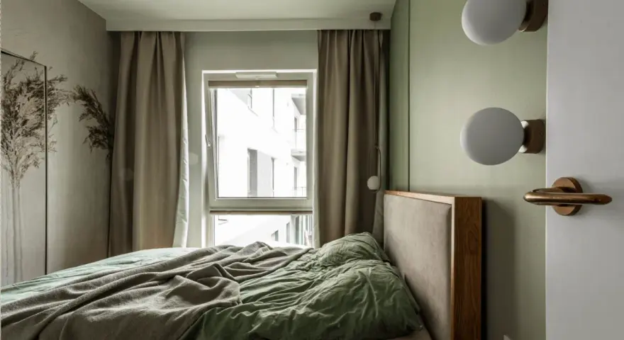 Przytulna, mała sypialnia z łóżkiem, zieloną pościelą i oknem z zasłonami. Dwie kule lamp na ścianie.
