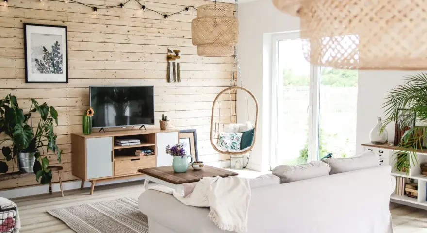 Przytulny salon z drewnianą ścianą, gdzie stara boazeria w nowej odsłonie tworzy ciepłą atmosferę. Wygodna sofa, wiszący fotel i rośliny dodają uroku.
