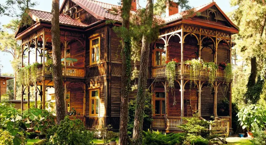 Drewniany dom w stylu świdermajer, z werandami i bogatymi zdobieniami, otoczony zielenią.