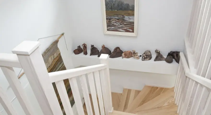 Jak wykończyć klatkę schodową w domu? Jasne drewno, białe balustrady i ciekawa ekspozycja rzeźb tworzą przytulną przestrzeń.