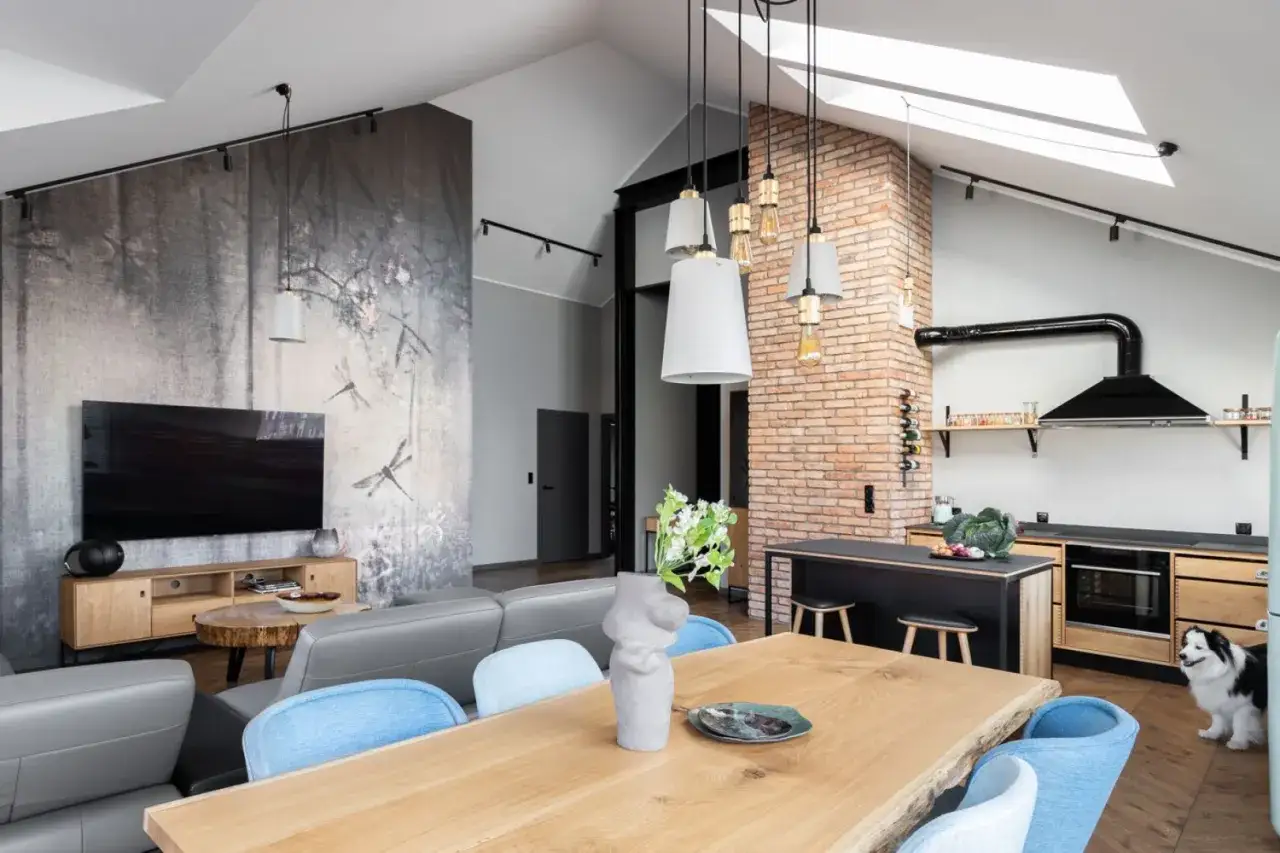 Nowoczesny salon loft z tapetą w ważki, szarą kanapą i drewnianym stołem. W tle ceglana ściana i kuchnia.