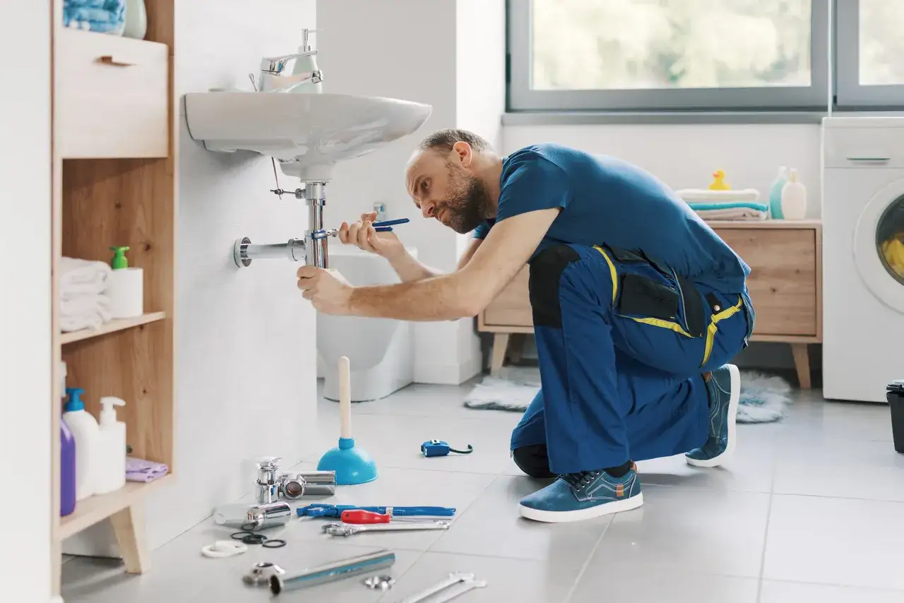 Instalator wykonuje montaż baterii nablatowej pod umywalką, używając klucza do dokręcenia syfonu.