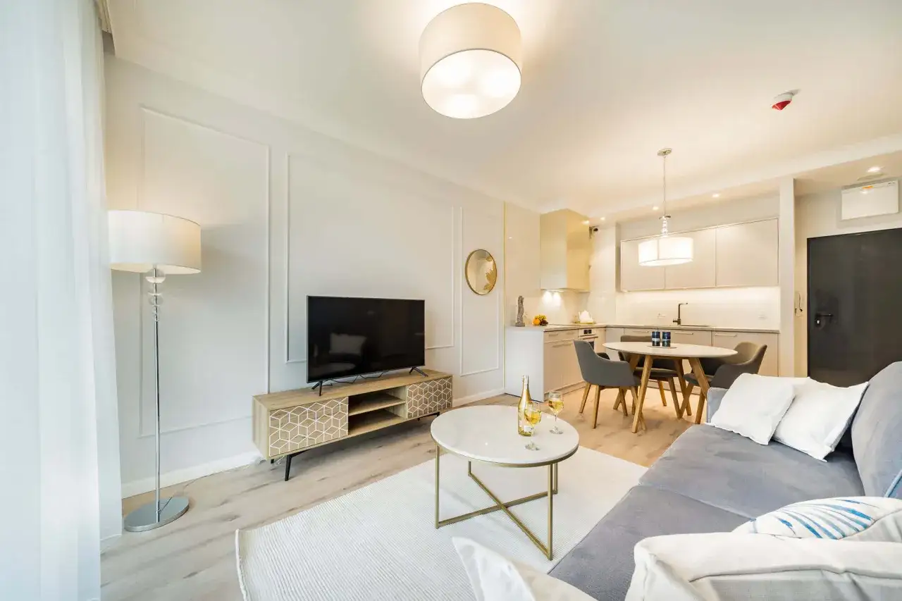 Elegancka sztukateria w nowoczesnym salonie z aneksem kuchennym. Jasne kolory, wygodna sofa i okrągły stolik tworzą przytulną atmosferę.